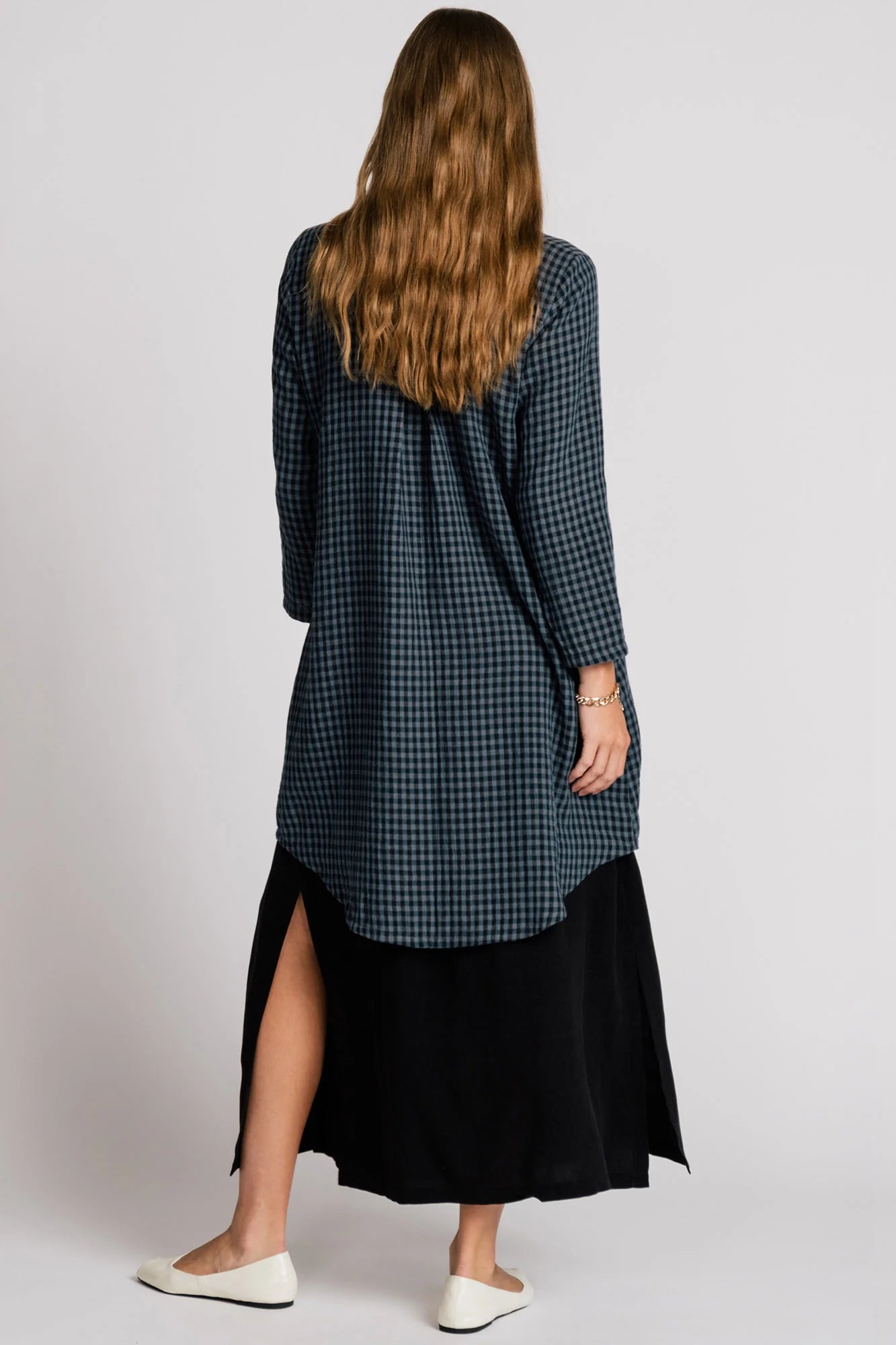 Back view of a woman wearing the Oriole Tunic by Allison Wonderland in Navy Gingham, a long tunic with a button front and rounded hi-low hemline. She is wearing it over a black skirt and standing against a grey background. 