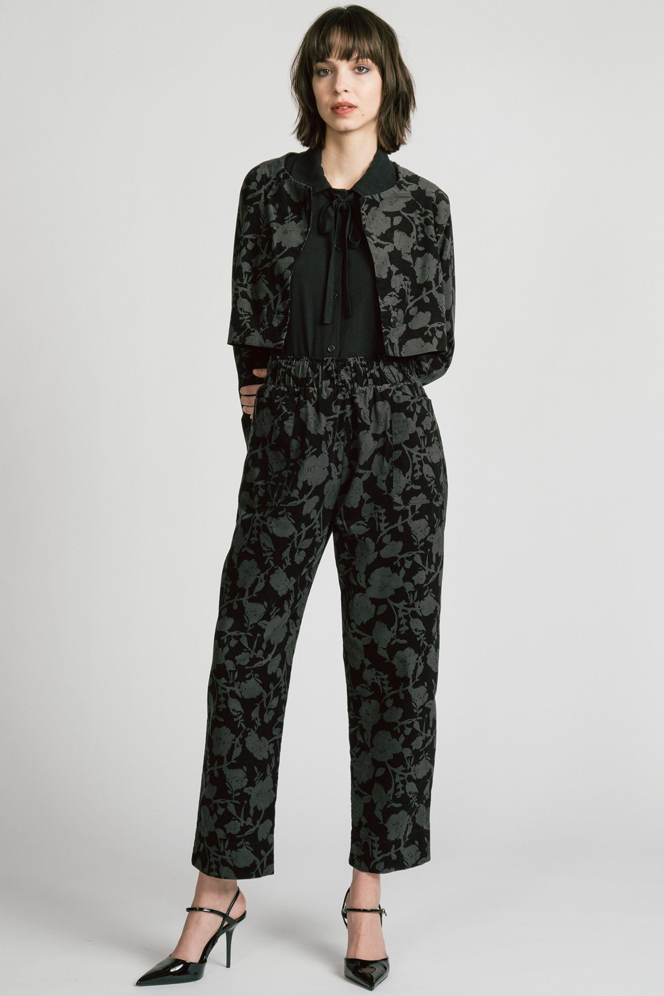 A woman wearing the Orion Pants by Allison Wonderland, relaxed fitting pants with an elastic waist and pockets in a dark Floral print on fine wale corduroy. She is wearing them with the matching Libra Bolero and standing in front of a white background. 
