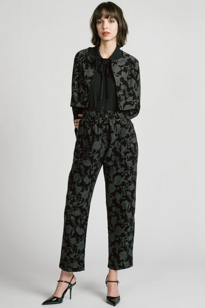 A woman wearing the Orion Pants by Allison Wonderland, relaxed fitting pants with an elastic waist and pockets in a dark Floral print on fine wale corduroy. She is wearing them with the matching Libra Bolero and standing in front of a white background. 