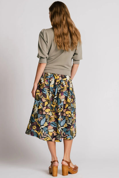 Back view of a woman wearing the Pelican skirt by Allison Wonderland in Black Floral, a mid-calf length tiered skirt with an elastic waist. She is wearing it with the Finch Sweatshirt in Eucalyptus and standing in front of a plain background. 