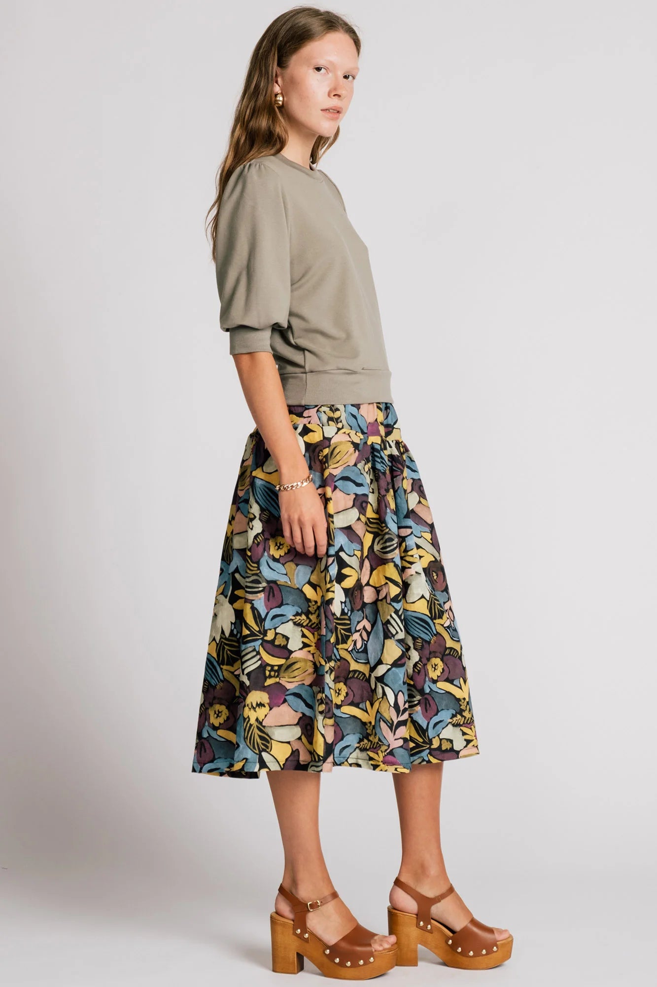 A woman wearing the Pelican skirt by Allison Wonderland in Black Floral, a mid-calf length tiered skirt with an elastic waist. She is wearing it with the Finch Sweatshirt in Eucalyptus and standing in front of a plain background. 