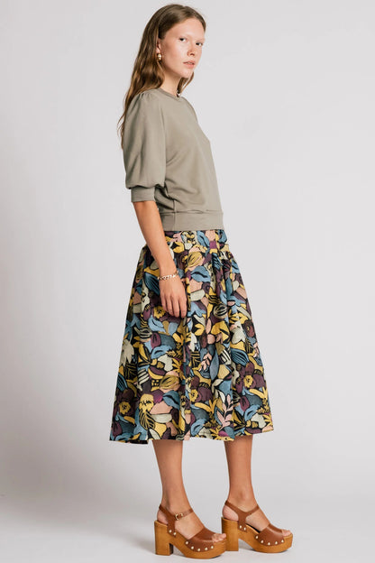 A woman wearing the Pelican skirt by Allison Wonderland in Black Floral, a mid-calf length tiered skirt with an elastic waist. She is wearing it with the Finch Sweatshirt in Eucalyptus and standing in front of a plain background. 