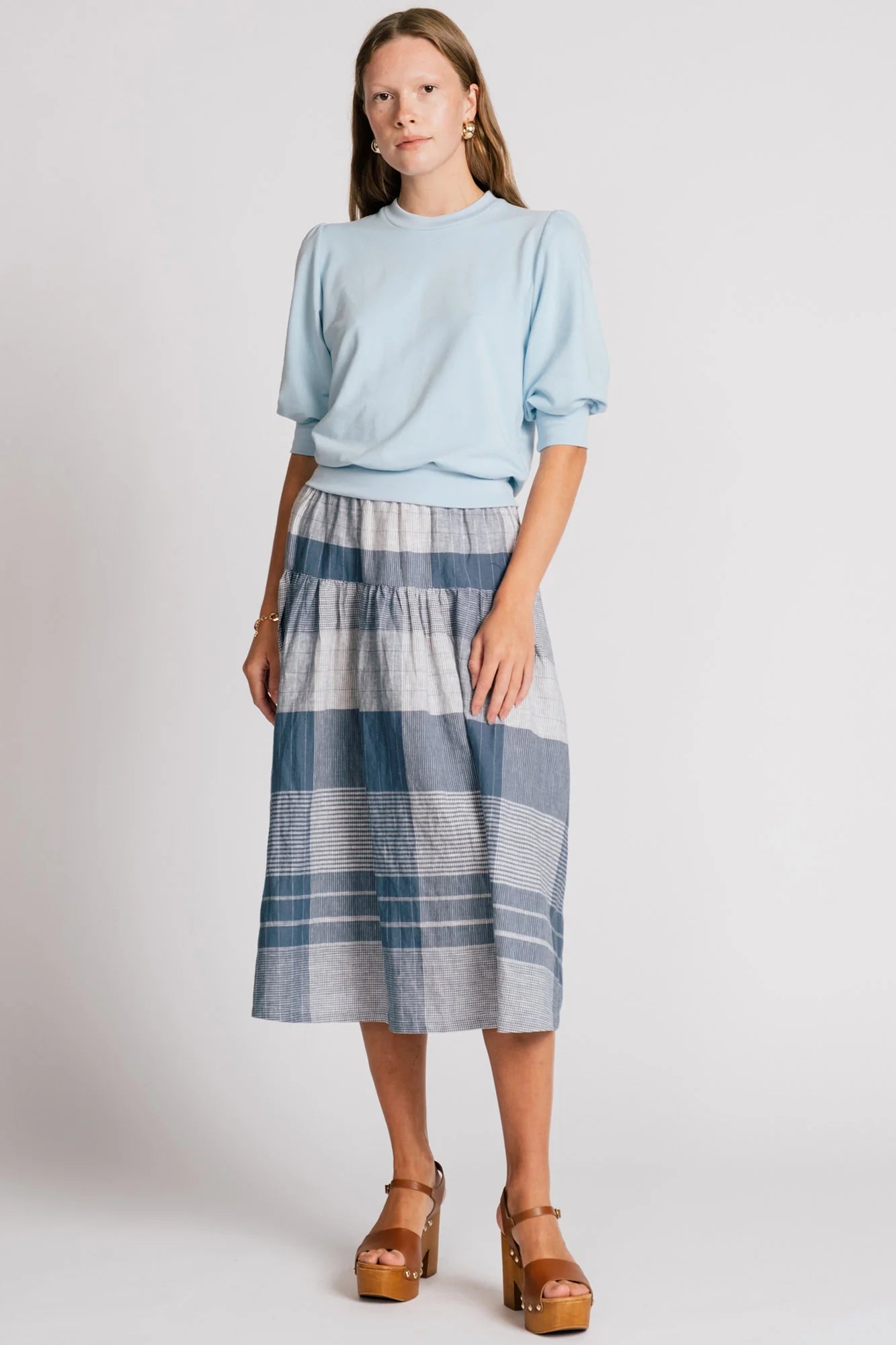 A woman wearing the Pelican skirt by Allison Wonderland in Blue Plaid, a mid-calf length tiered skirt with an elastic waist. She is wearing it with the Finch Sweatshirt in Baby Blue and standing in front of a plain background. 