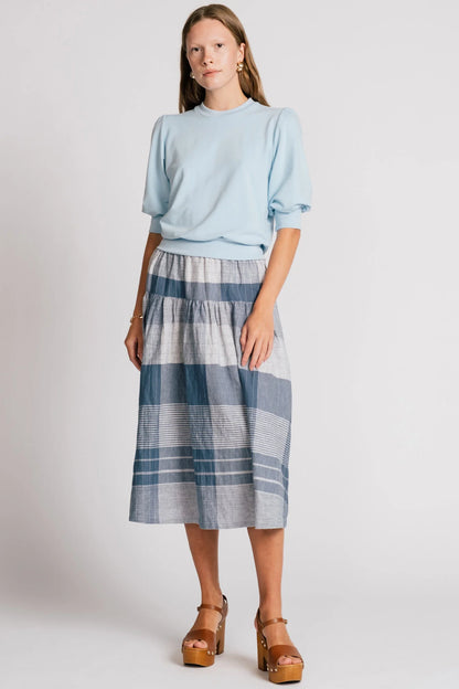 A woman wearing the Pelican skirt by Allison Wonderland in Blue Plaid, a mid-calf length tiered skirt with an elastic waist. She is wearing it with the Finch Sweatshirt in Baby Blue and standing in front of a plain background. 