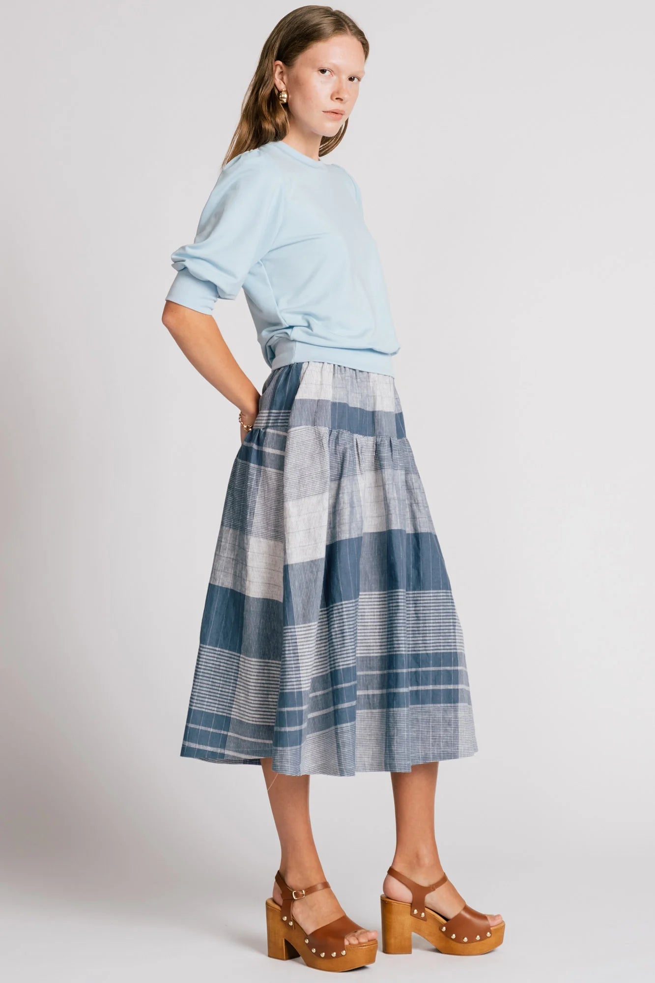 A woman wearing the Pelican skirt by Allison Wonderland in Blue Plaid, a mid-calf length tiered skirt with an elastic waist. She is wearing it with the Finch Sweatshirt in Baby Blue and standing in front of a plain background. 
