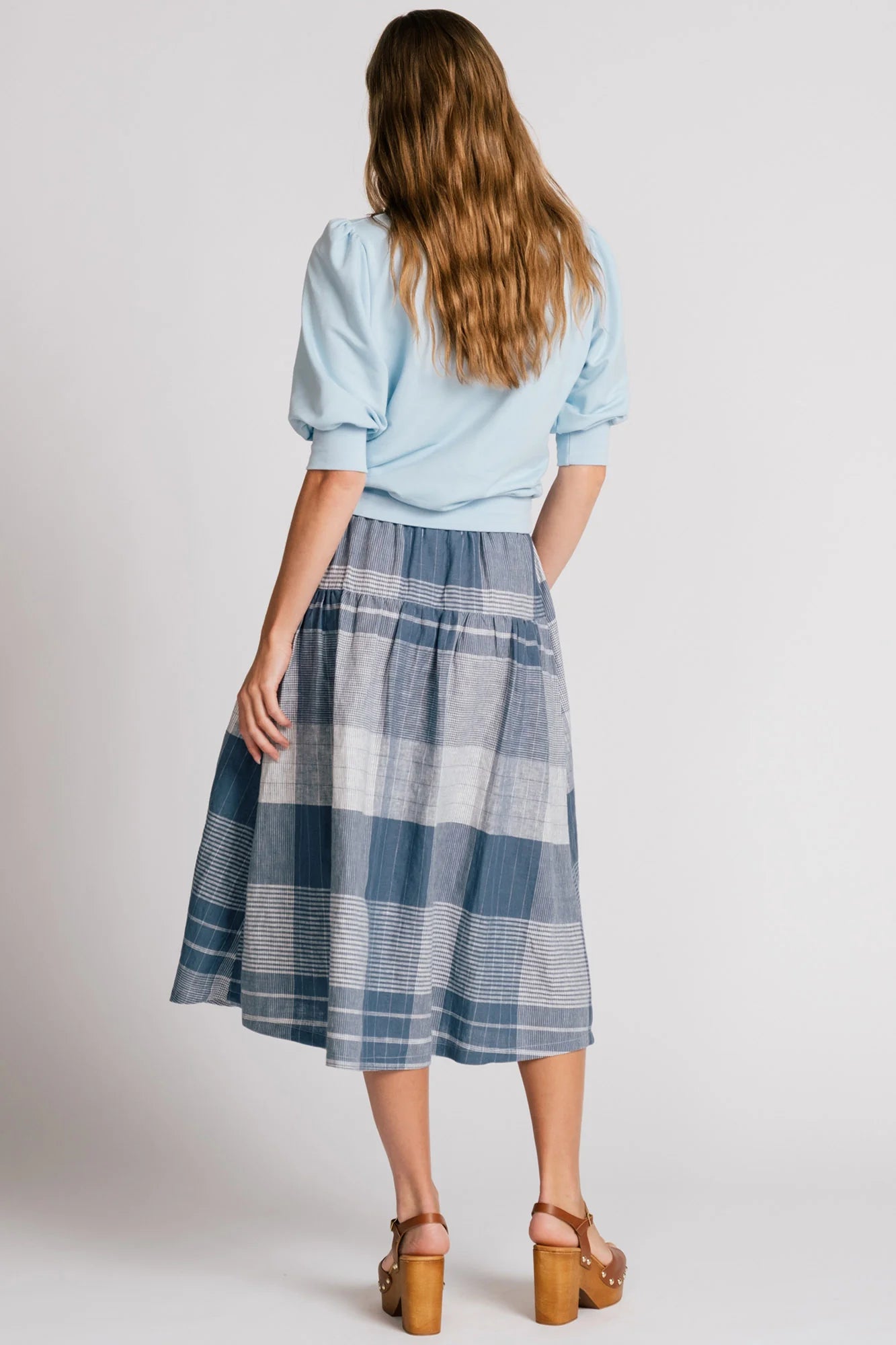 Back view of a woman wearing the Pelican skirt by Allison Wonderland in Blue Plaid, a mid-calf length tiered skirt with an elastic waist. She is wearing it with the Finch Sweatshirt in Baby Blue and standing in front of a plain background. 