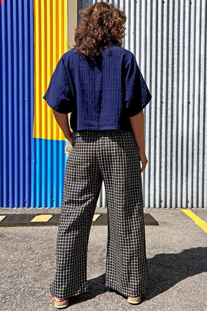 Back view of a woman wearing the Pip Jacket by Allison Wonderland in Navy/Black Stripe, with 
checkered pants standing against a colourful wall.