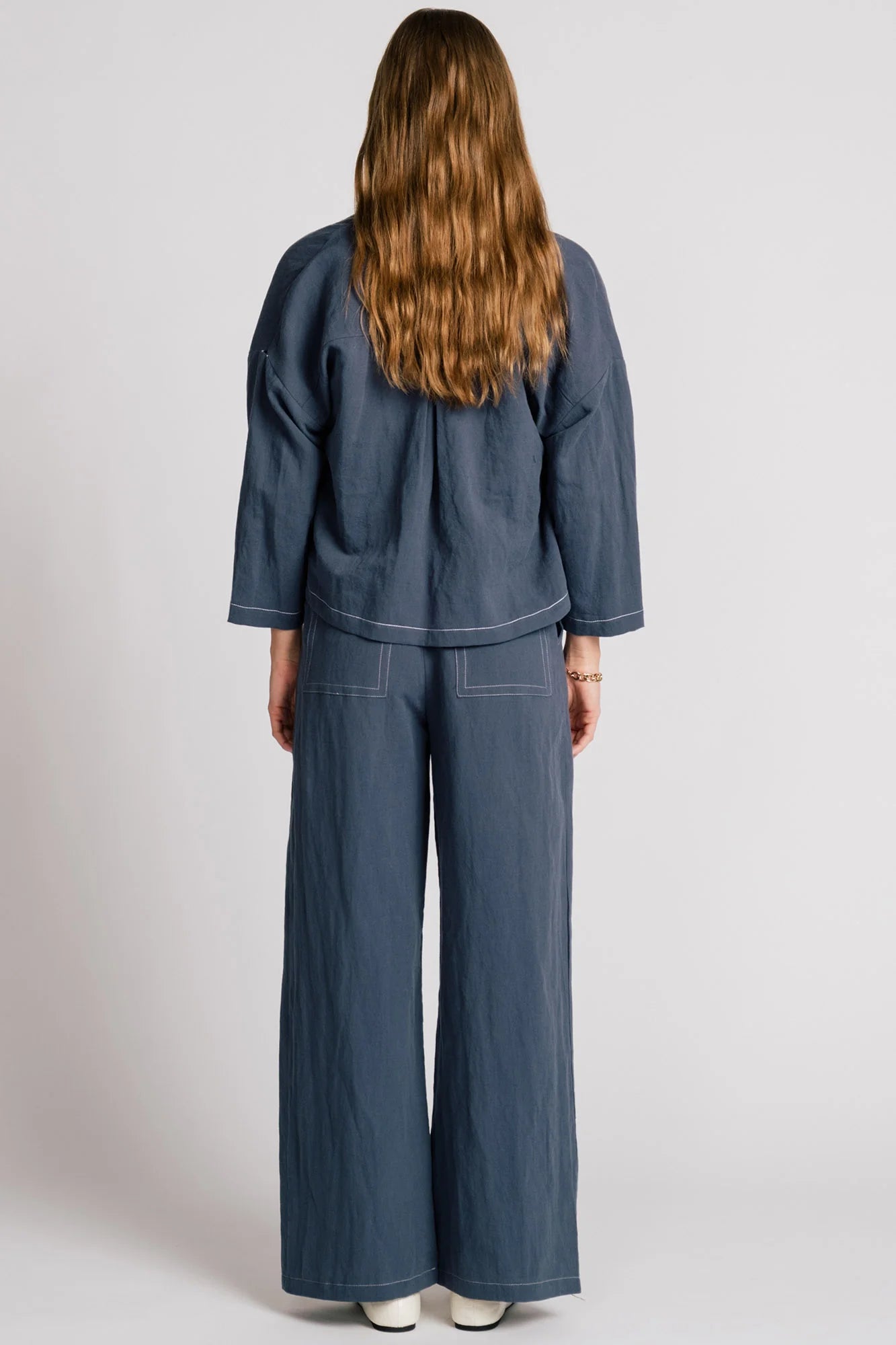 Back view of a woman wearing the Tern Blazer by Allison Wonderland in Navy, a cropped open jacket with a boxy fit and two front patch pockets. She is wearing it with the matching Whistler Pants and a gingham top and standing against a white background. 