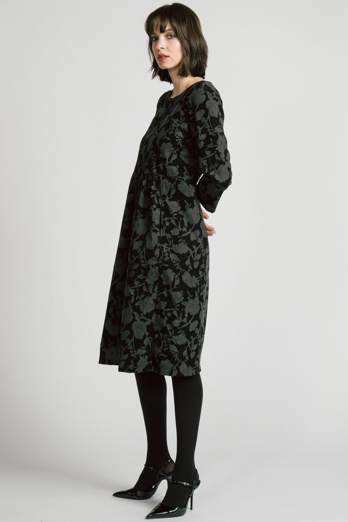 Side view of a woman wearing the Virgo Dress by Allison Wonderland in Floral, a babydoll style dress in floral printed fine wale corduroy with bracelet length sleeves and a midi length. She's standing against a white background. 