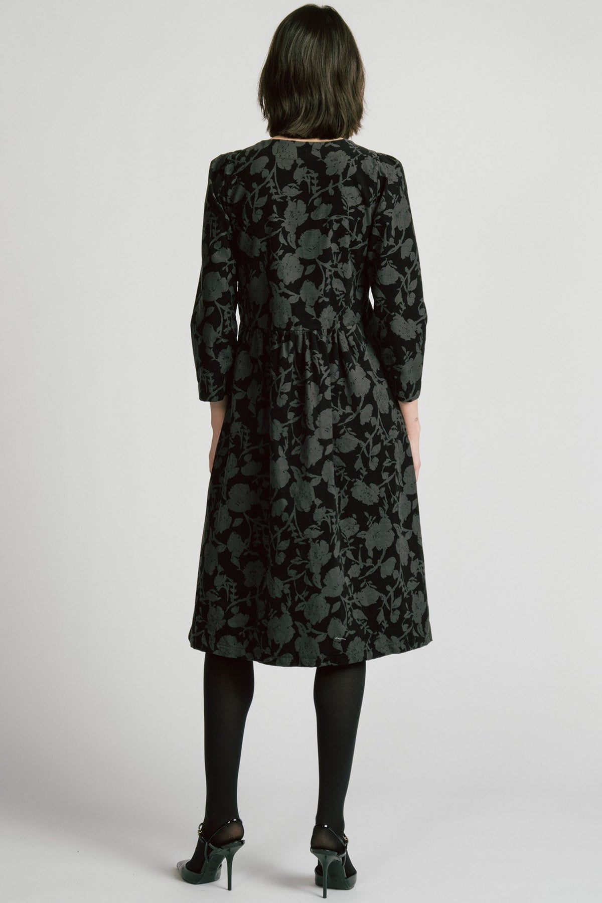 Back view of a woman wearing the Virgo Dress by Allison Wonderland in Floral, a babydoll style dress in floral printed fine wale corduroy with bracelet length sleeves and a midi length. She's standing against a white background. 