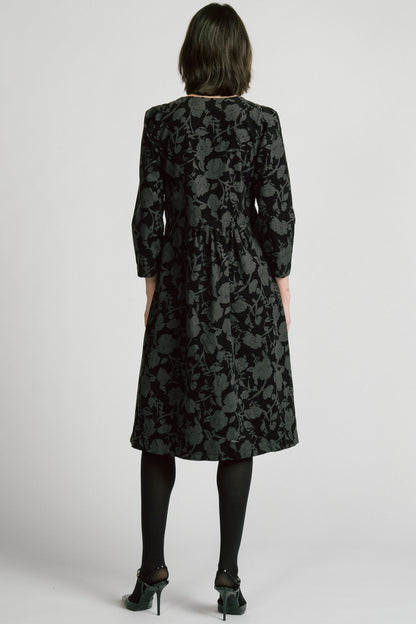 Back view of a woman wearing the Virgo Dress by Allison Wonderland in Floral, a babydoll style dress in floral printed fine wale corduroy with bracelet length sleeves and a midi length. She's standing against a white background. 