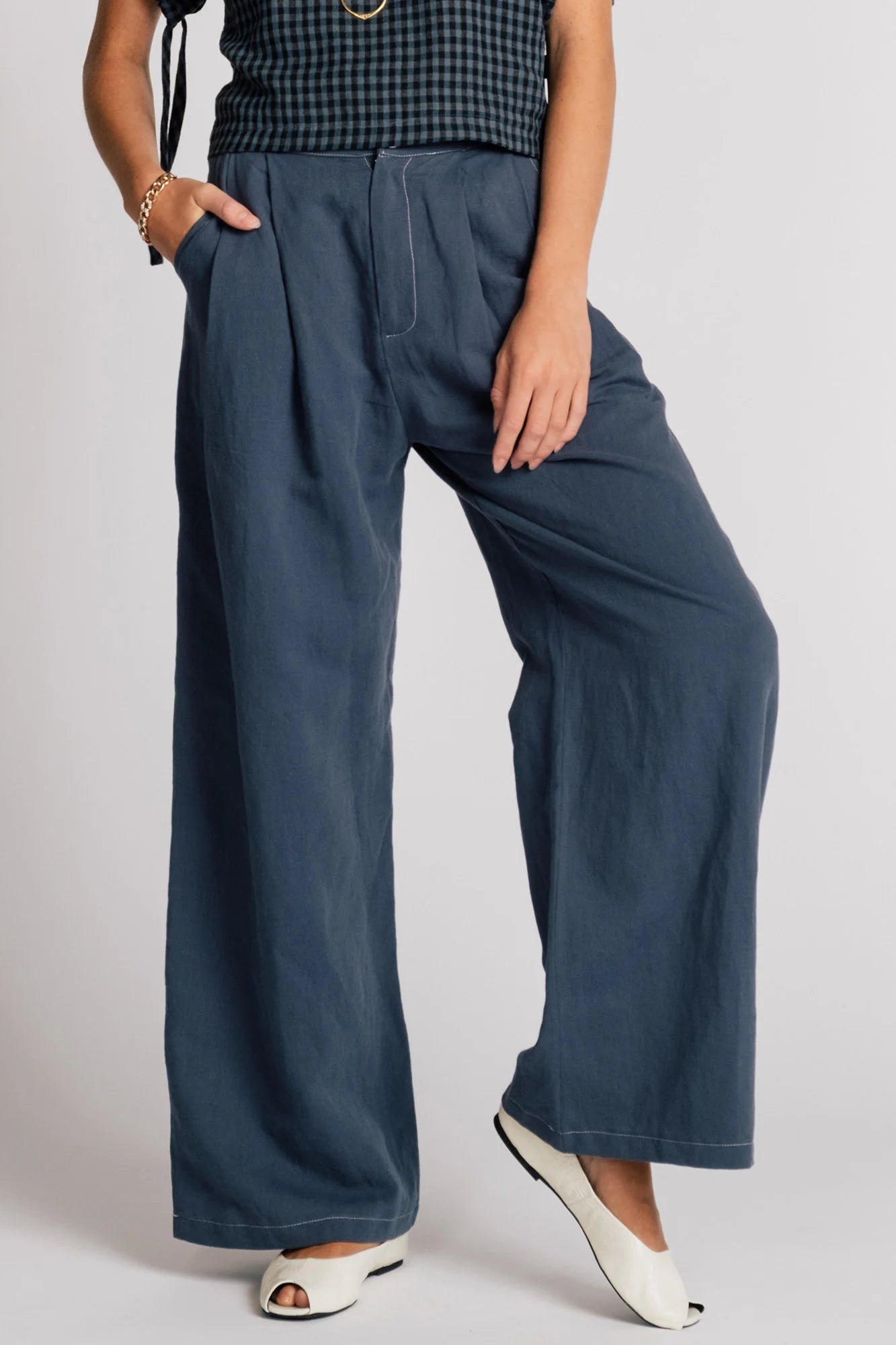 Waist down view of a woman wearing the Whistler Pants by Allison Wonderland in Navy, wide legged pants with front pleats, a zip fly, and contrast white stitching. She is wearing them with a dark gingham top and standing in front of a white background. 