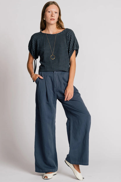 A woman wearing the Whistler Pants by Allison Wonderland in Navy, wide legged pants with front pleats, a zip fly, and contrast white stitching. She is wearing them with a dark gingham top and standing in front of a white background. 