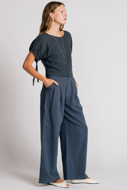 A woman wearing the Whistler Pants by Allison Wonderland in Navy, wide legged pants with front pleats, a zip fly, and contrast white stitching. She is wearing them with a dark gingham top and standing in front of a white background. 