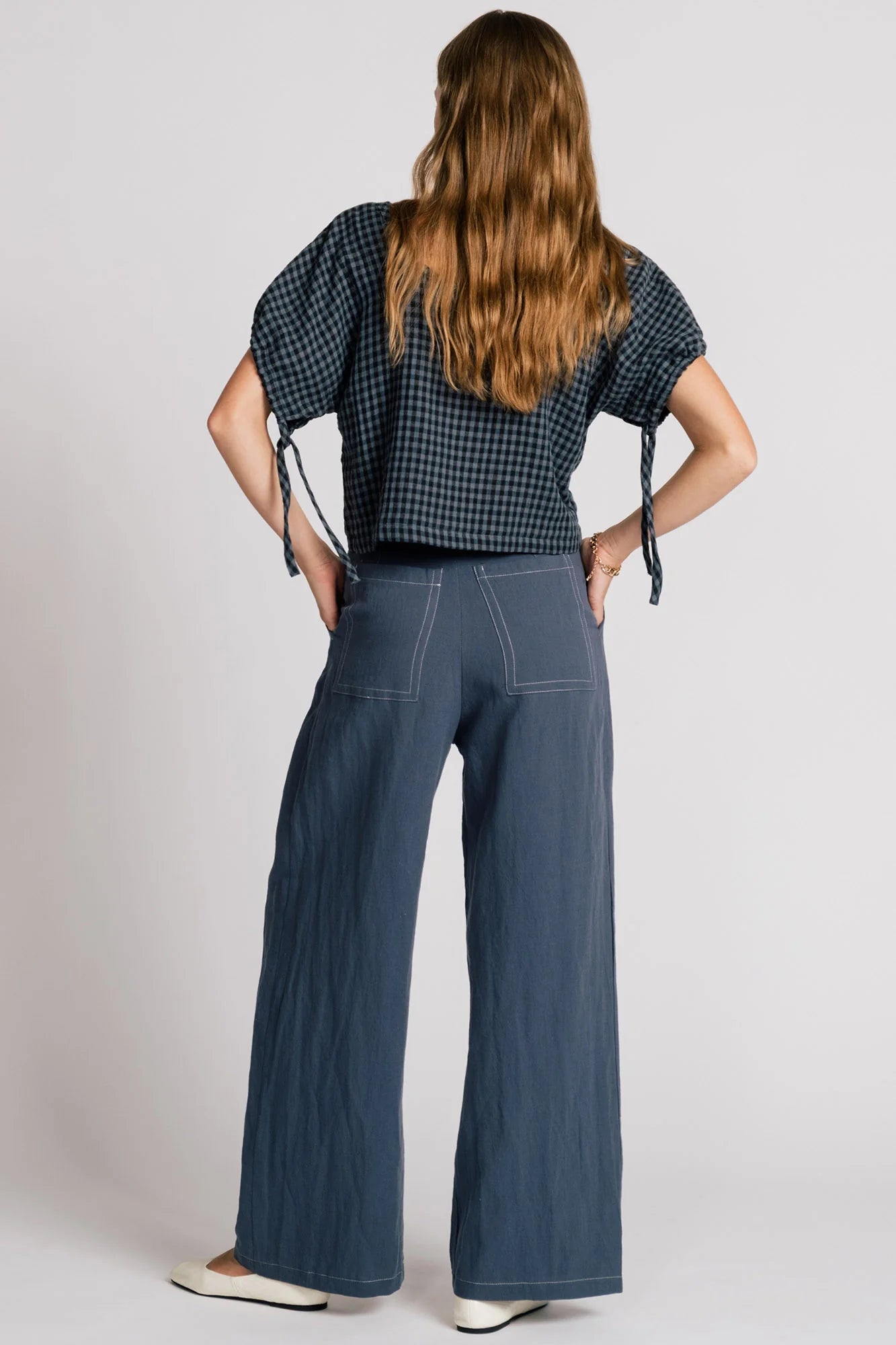 Back view of a woman wearing the Whistler Pants by Allison Wonderland in Navy, wide legged pants with front pleats, a zip fly, and contrast white stitching. She is wearing them with a dark gingham top and standing in front of a white background. 