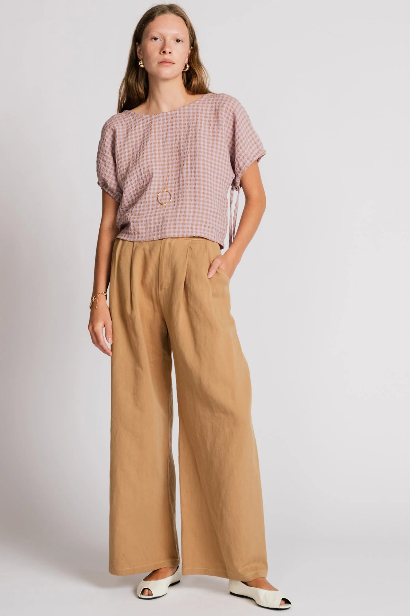 A woman wearing the Whistler Pants by Allison Wonderland in Sand, wide legged pants with front pleats, a zip fly, and contrast white stitching. She is wearing them with a light gingham top and standing in front of a white background. 