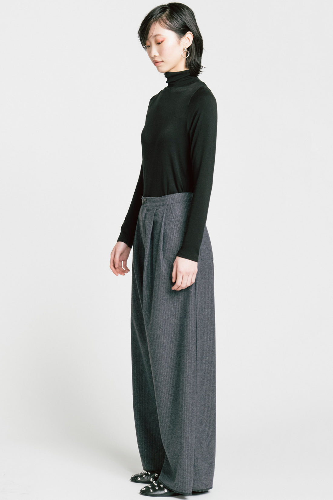 A woman wearing the Aches Turtleneck by Allison Wonderland in Black, a slim fitting classic turtleneck. She is wearing it with the Memory Pants in Denim Pinstripe and standing in front of a white background. 