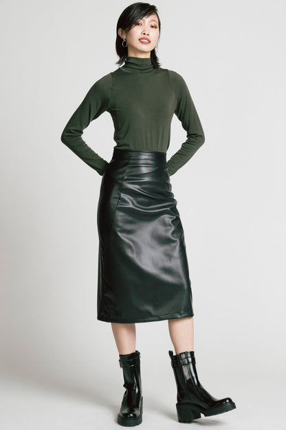 A woman wearing the Aches Turtleneck by Allison Wonderland in Green, a slim fitting classic turtleneck. She is wearing it with the Desire Skirt in Black and standing in front of a white background. 