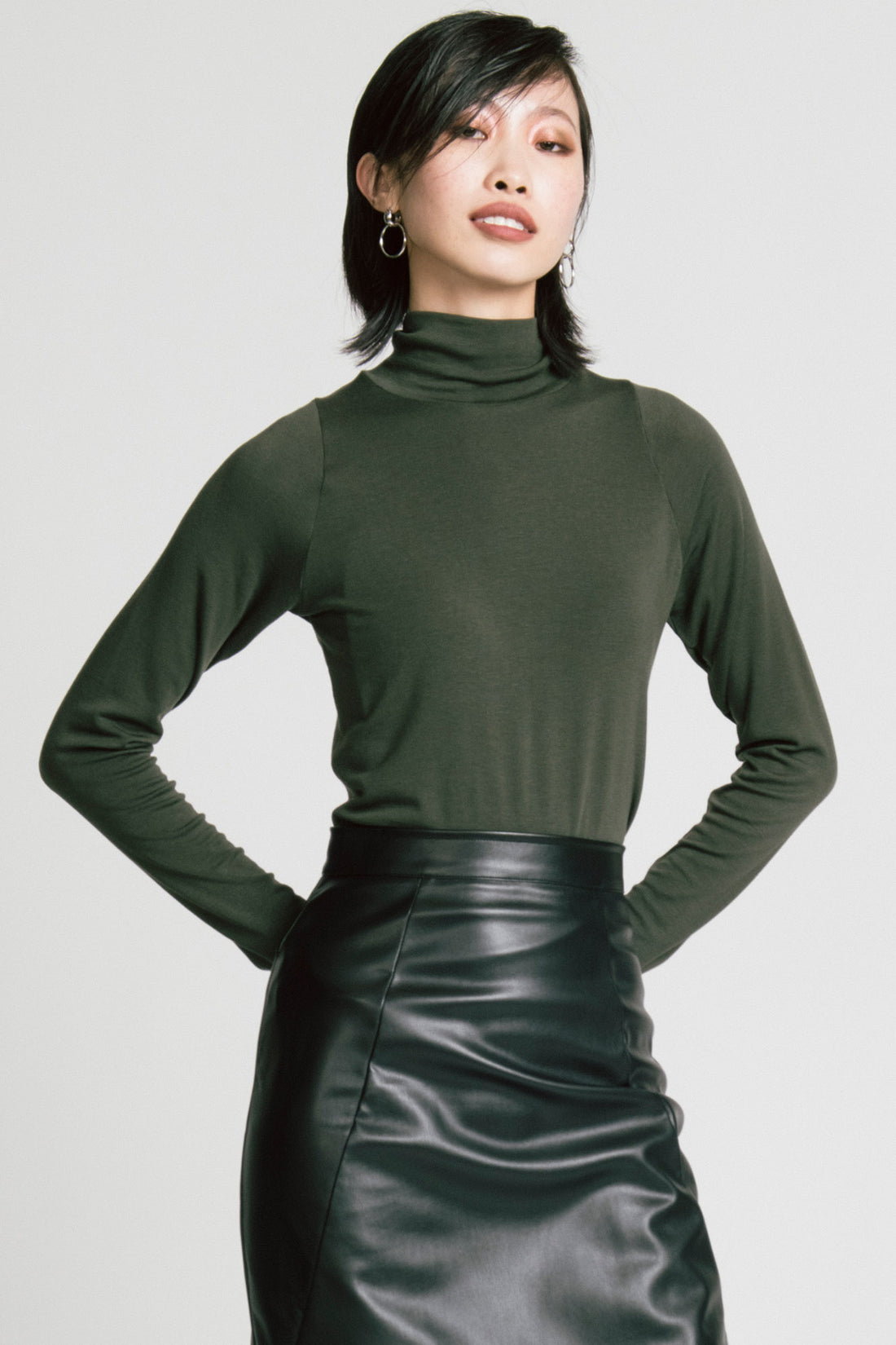 A woman wearing the Aches Turtleneck by Allison Wonderland in Green, a slim fitting classic turtleneck. She is wearing it with the Desire Skirt in Black and standing in front of a white background. 