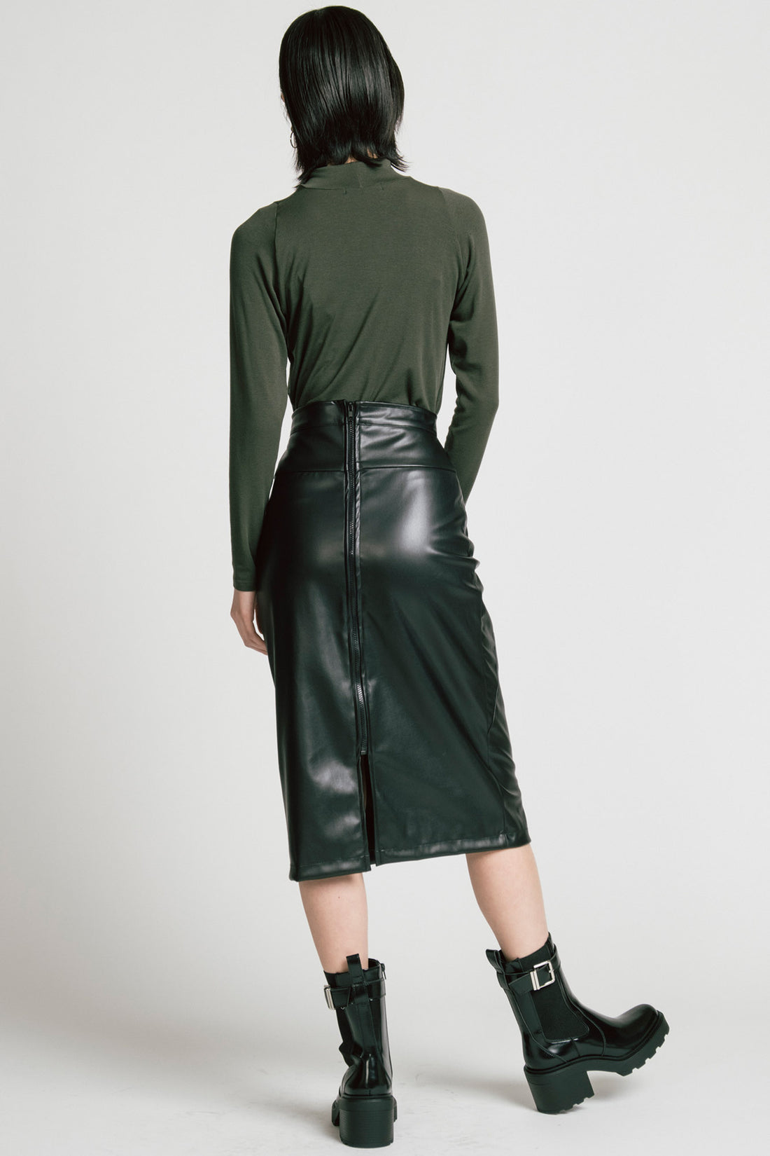 Back view of a  woman wearing the Aches Turtleneck by Allison Wonderland in Green, a slim fitting classic turtleneck. She is wearing it with the Desire Skirt in Black and standing in front of a white background. 