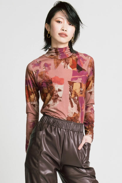 A woman wearing the Aches Turtleneck by Allison Wonderland in Burgundy print, a slim fitting classic turtleneck in an abstract pattern. She is wearing it with brown faux leather pants and standing in front of a white background. 