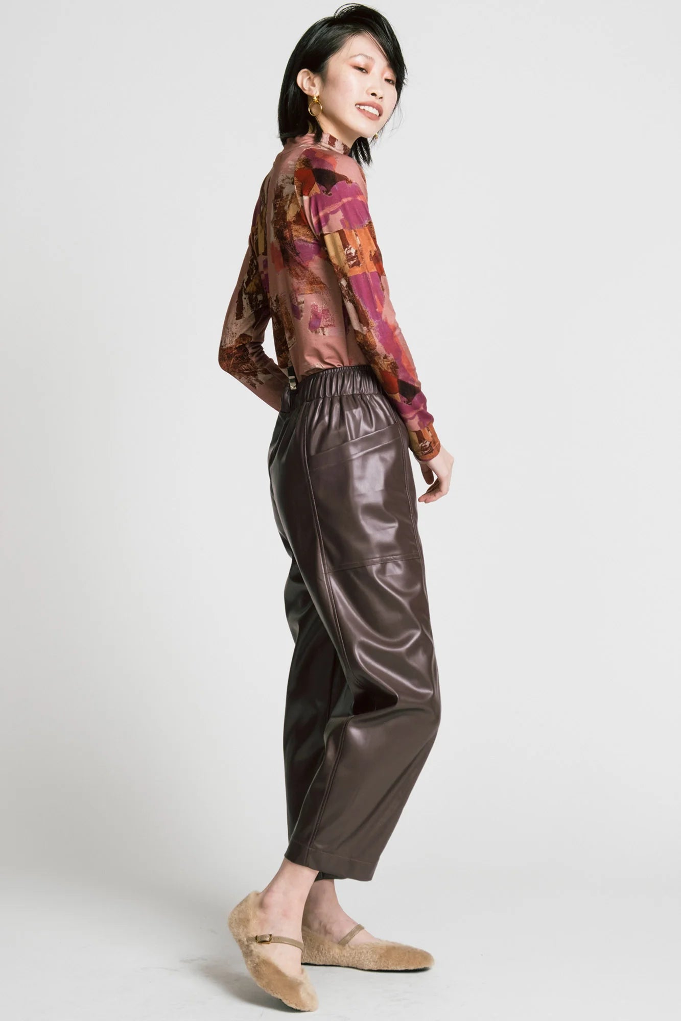 Side view of a woman wearing the Aches Turtleneck by Allison Wonderland in Burgundy print, a slim fitting classic turtleneck in an abstract pattern. She is wearing it with brown faux leather pants and standing in front of a white background. 
