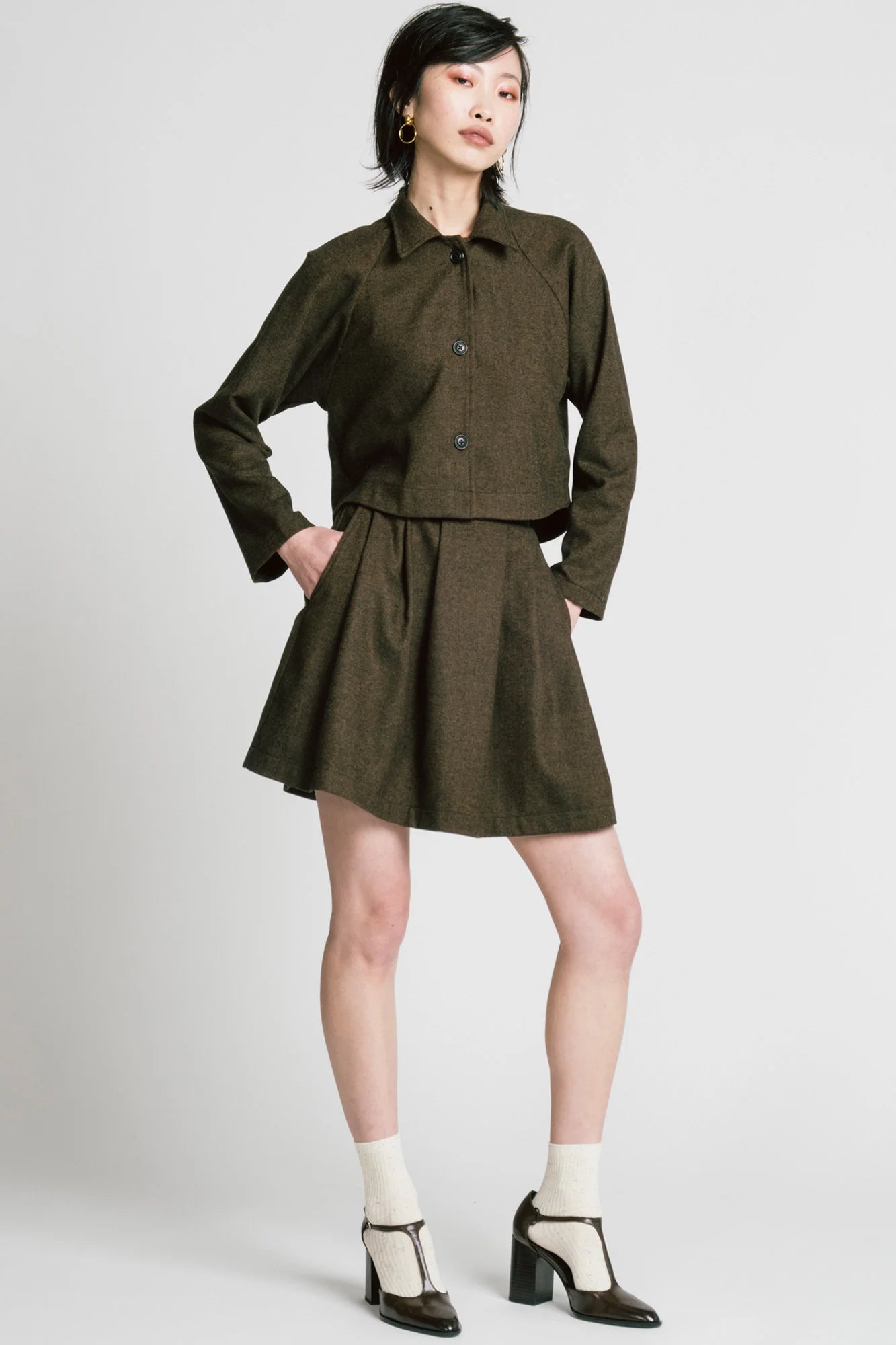 A woman wearing the Cycle Jacket by Allison Wonderland in brown Herringbone, a cropped jacket with a lapel collar, button front, and raglan sleeves. She is wearing it with a matching skirt and standing in front of a white background. 
