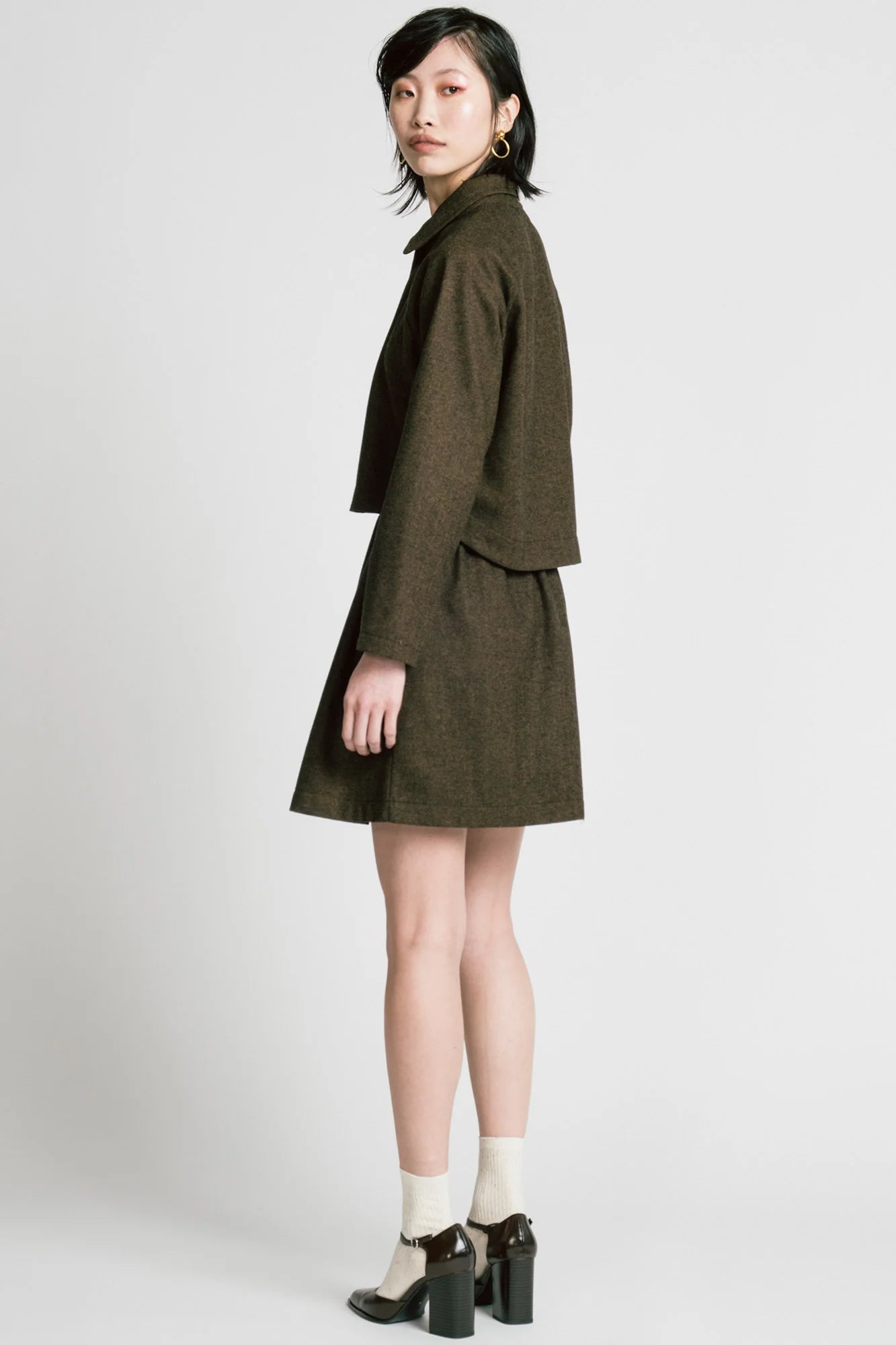 Side view of a woman wearing the Cycle Jacket by Allison Wonderland in brown Herringbone, a cropped jacket with a lapel collar, button front, and raglan sleeves. She is wearing it with a matching skirt and standing in front of a white background. 