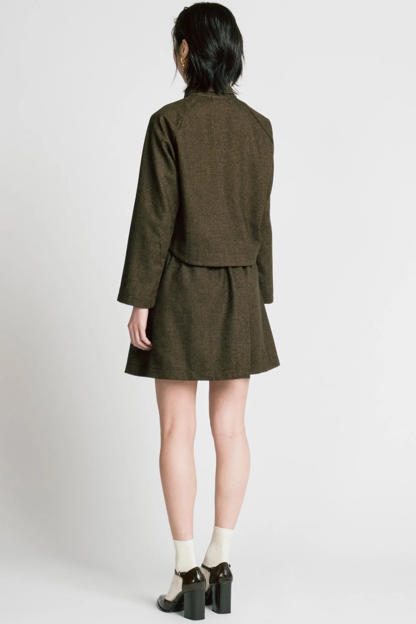 Back view of a woman wearing the Cycle Jacket by Allison Wonderland in brown Herringbone, a cropped jacket with a lapel collar, button front, and raglan sleeves. She is wearing it with a matching skirt and standing in front of a white background. 