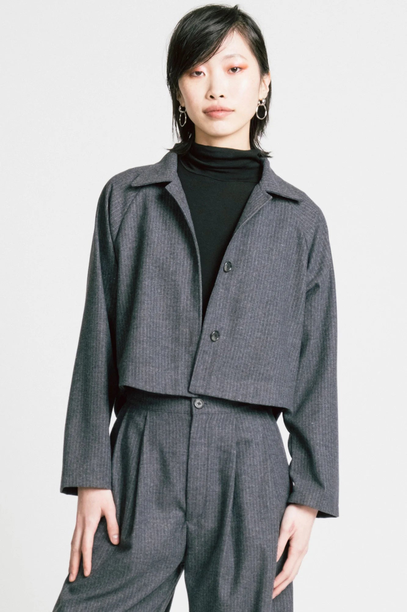 3/4 view of a woman wearing the Cycle Jacket by Allison Wonderland in denim Pinstripe, a cropped jacket with a lapel collar, button front, and raglan sleeves. She is wearing it with the a black turtleneck and the matching Memory Pants and standing in front of a white background. 