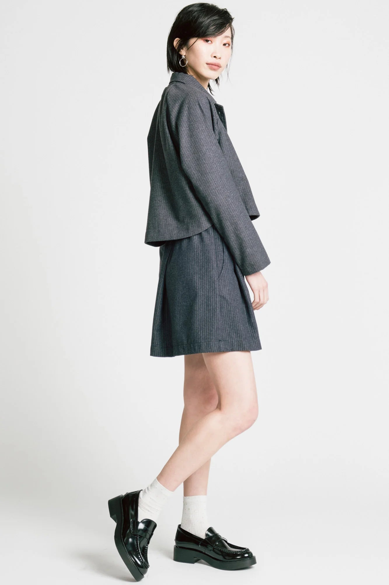Side view of a woman wearing the Cycle Jacket by Allison Wonderland in denim Pinstripe, a cropped jacket with a lapel collar, button front, and raglan sleeves. She is wearing it with a matching skirt and standing in front of a white background. 