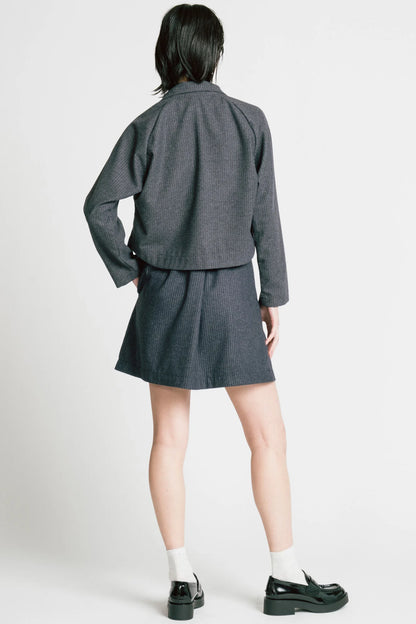 Back view of a woman wearing the Cycle Jacket by Allison Wonderland in denim Pinstripe, a cropped jacket with a lapel collar, button front, and raglan sleeves. She is wearing it with a matching skirt and standing in front of a white background. 
