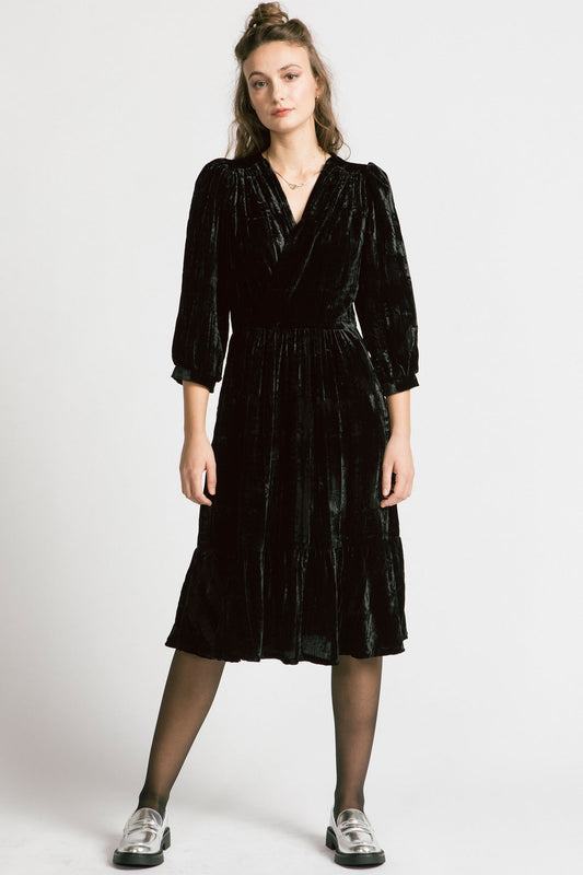 A woman wearing the Dark Lover Dress by Allison Wonderland in Black crushed velvet, featuring a faux-wrap front, 3/4 gathered sleeves, and a tiered midi-length skirt. She is standing in front of a white background. 