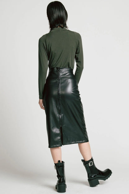 Back view of a woman wearing the Desire Skirt by Allison Wonderland in Black faux leather, a midi-length pencil skirt with a two-way back zipper. She is wearing it with a green turtleneck and standing in front of a white background. 