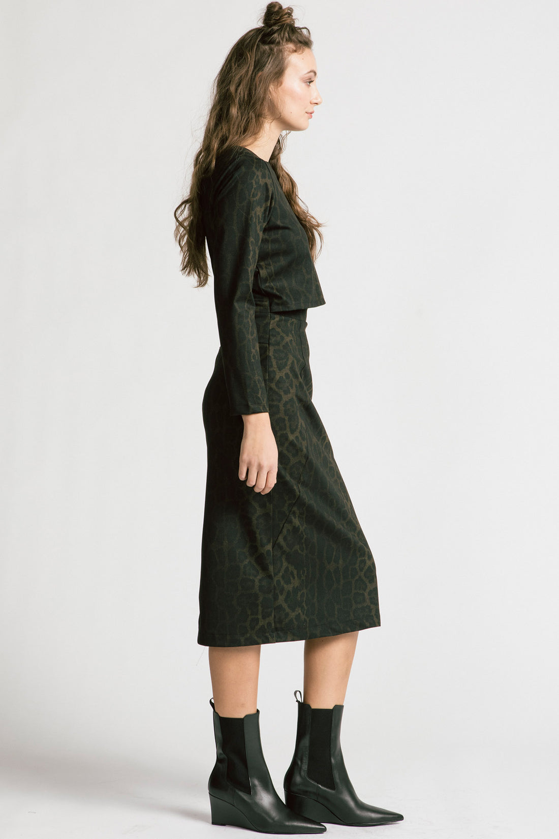 Side view of a woman wearing the Allison Wonderland Expectations dress in Leopard, featuring an opening at the front and attached back, a two-way back zipper, V-neckline at the back, long sleeves, and midi-length. She is standing in front of a white background. 