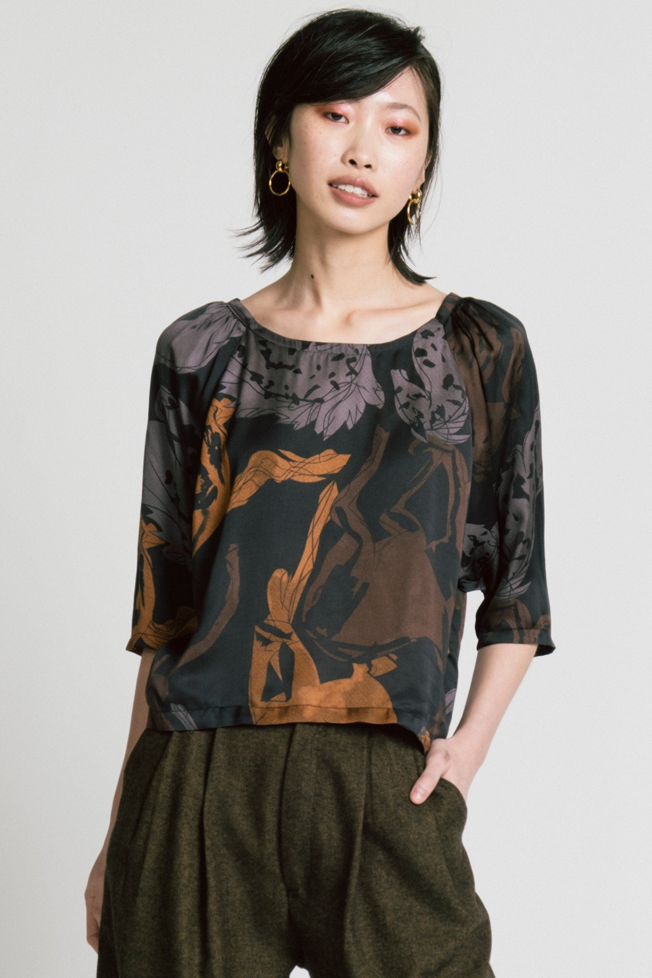 3/4 view of a woman wearing the Flash Blouse by Allison Wonderland in Horse Print, featuring 3/4 dropped raglan sleeves, a boat neck with gathers at the shoulders, and a loose fit. She is wearing it with the Memory Pant in Herringbone and standing in front of a white background. 