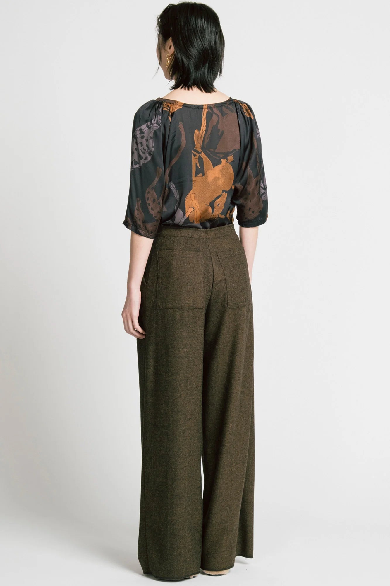 Back view of a woman wearing the Flash Blouse by Allison Wonderland in Horse Print, featuring 3/4 dropped raglan sleeves, a boat neck with gathers at the shoulders, and a loose fit. She is wearing it with the Memory Pant in Herringbone and standing in front of a white background. 