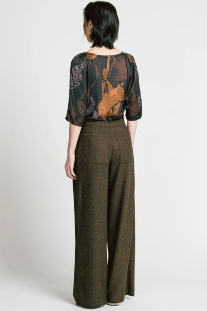 Back view of a woman wearing the Flash Blouse by Allison Wonderland in Horse Print, featuring 3/4 dropped raglan sleeves, a boat neck with gathers at the shoulders, and a loose fit. She is wearing it with the Memory Pant in Herringbone and standing in front of a white background. 