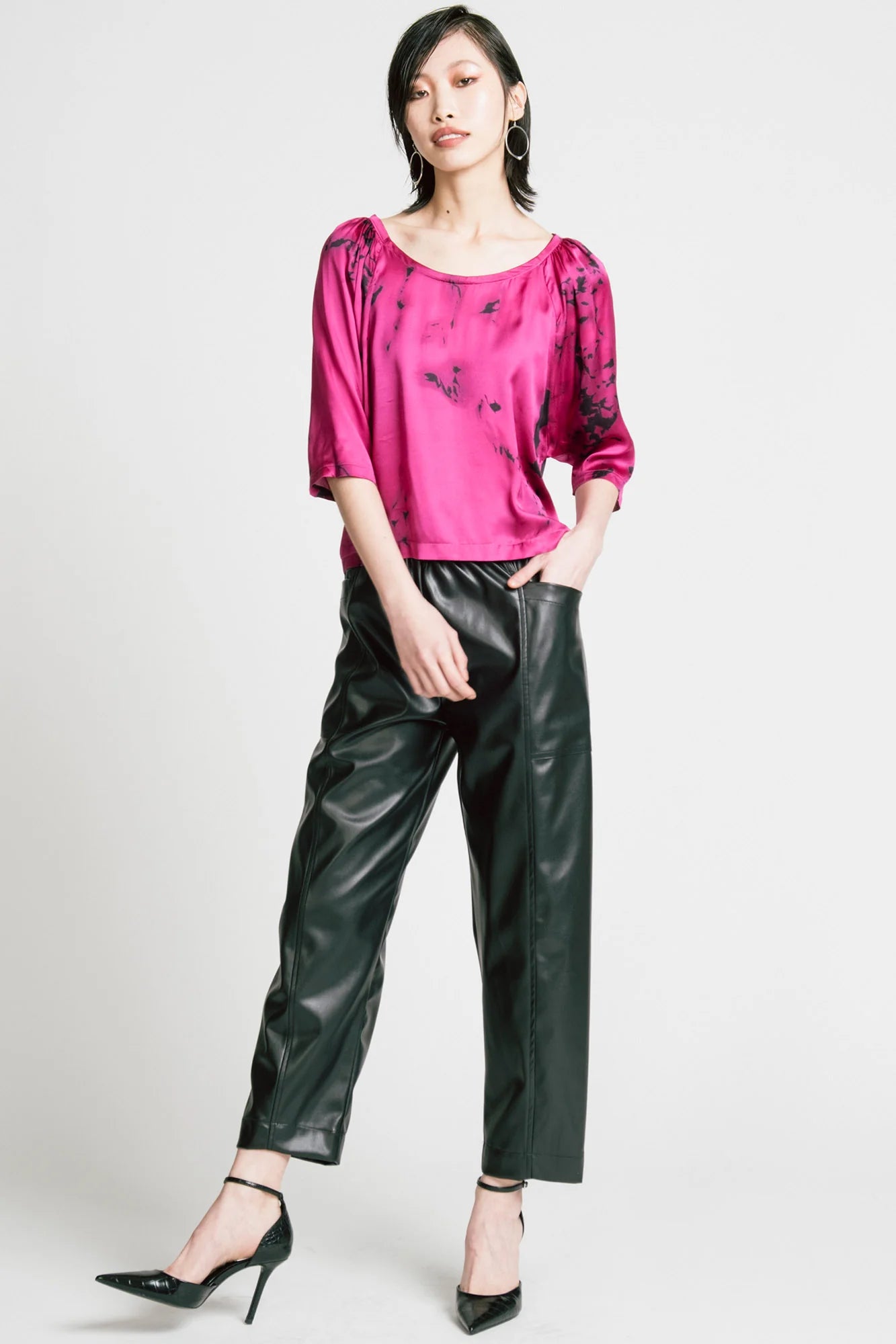 A woman wearing the Flash Blouse by Allison Wonderland in Pink Print, featuring 3/4 dropped raglan sleeves, a boat neck with gathers at the shoulders, and a loose fit. She is wearing it with black faux leather pants and standing in front of a white background.