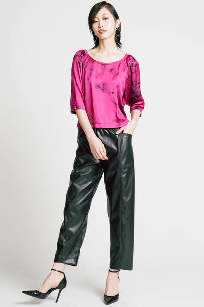 A woman wearing the Flash Blouse by Allison Wonderland in Pink Print, featuring 3/4 dropped raglan sleeves, a boat neck with gathers at the shoulders, and a loose fit. She is wearing it with black faux leather pants and standing in front of a white background.