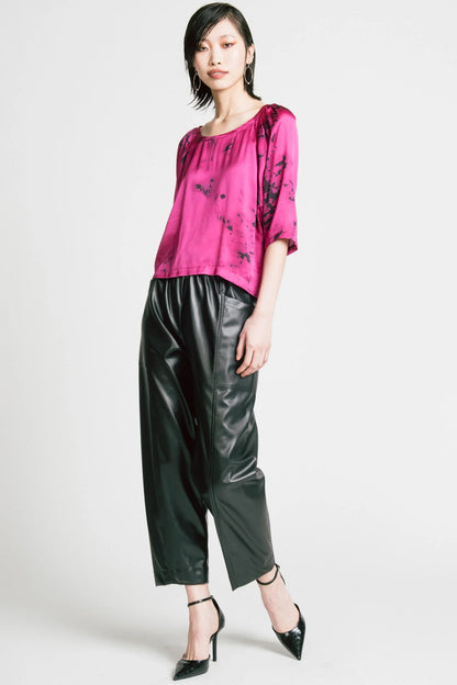 A woman wearing the Flash Blouse by Allison Wonderland in Pink Print, featuring 3/4 dropped raglan sleeves, a boat neck with gathers at the shoulders, and a loose fit. She is wearing it with black faux leather pants and standing in front of a white background.