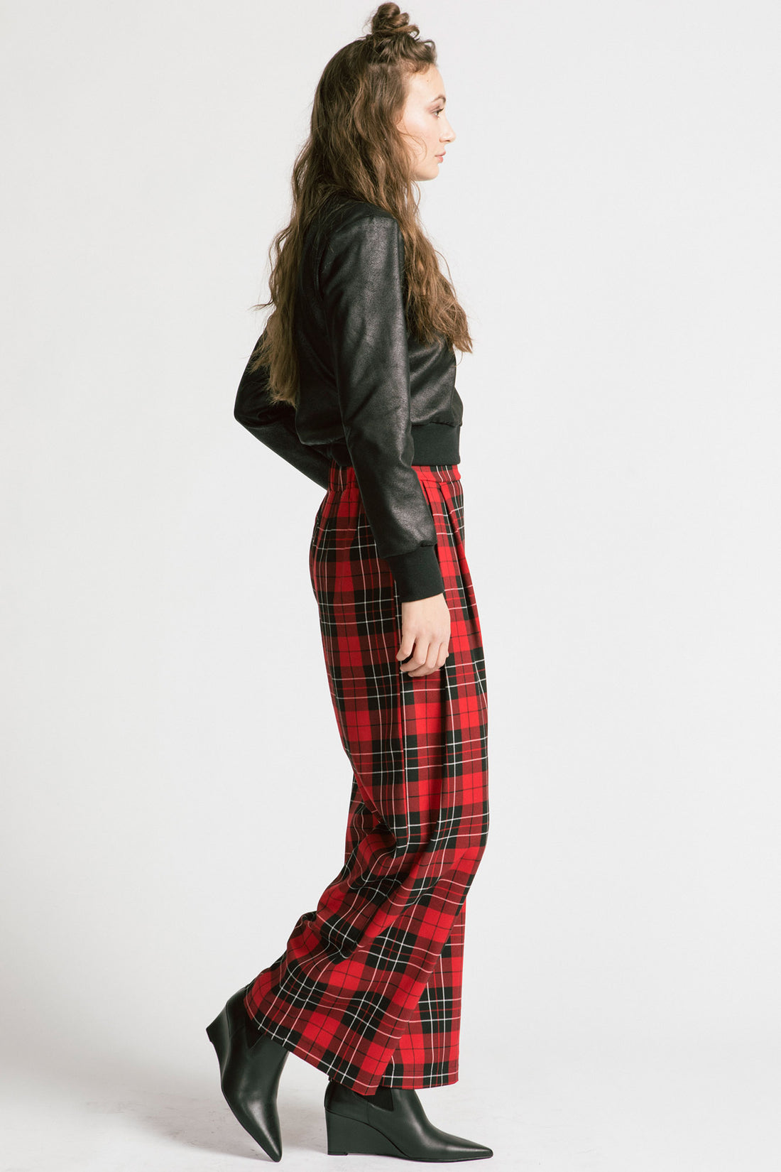 Side view of a woman wearing the Gone Top by Allison Wonderland in Leatherette, a classic crewneck in faux leather fabric, with red plaid pants. She is standing in front of a white background 