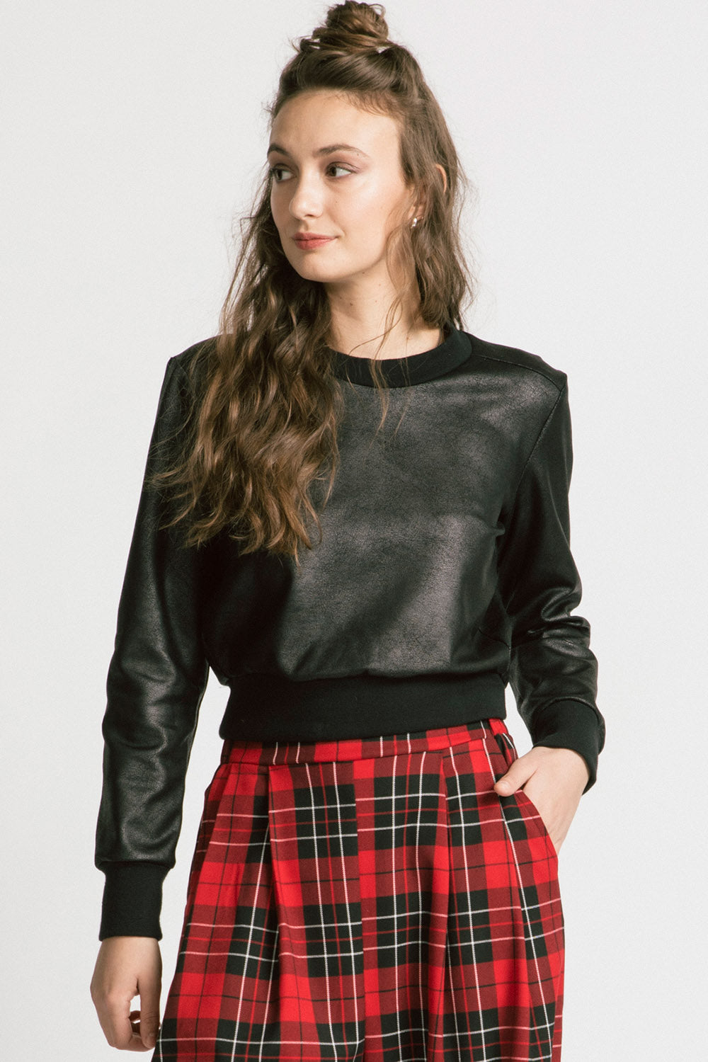 A woman wearing the Gone Top by Allison Wonderland in Leatherette, a classic crewneck in faux leather fabric, with red plaid pants. She is standing in front of a white background 