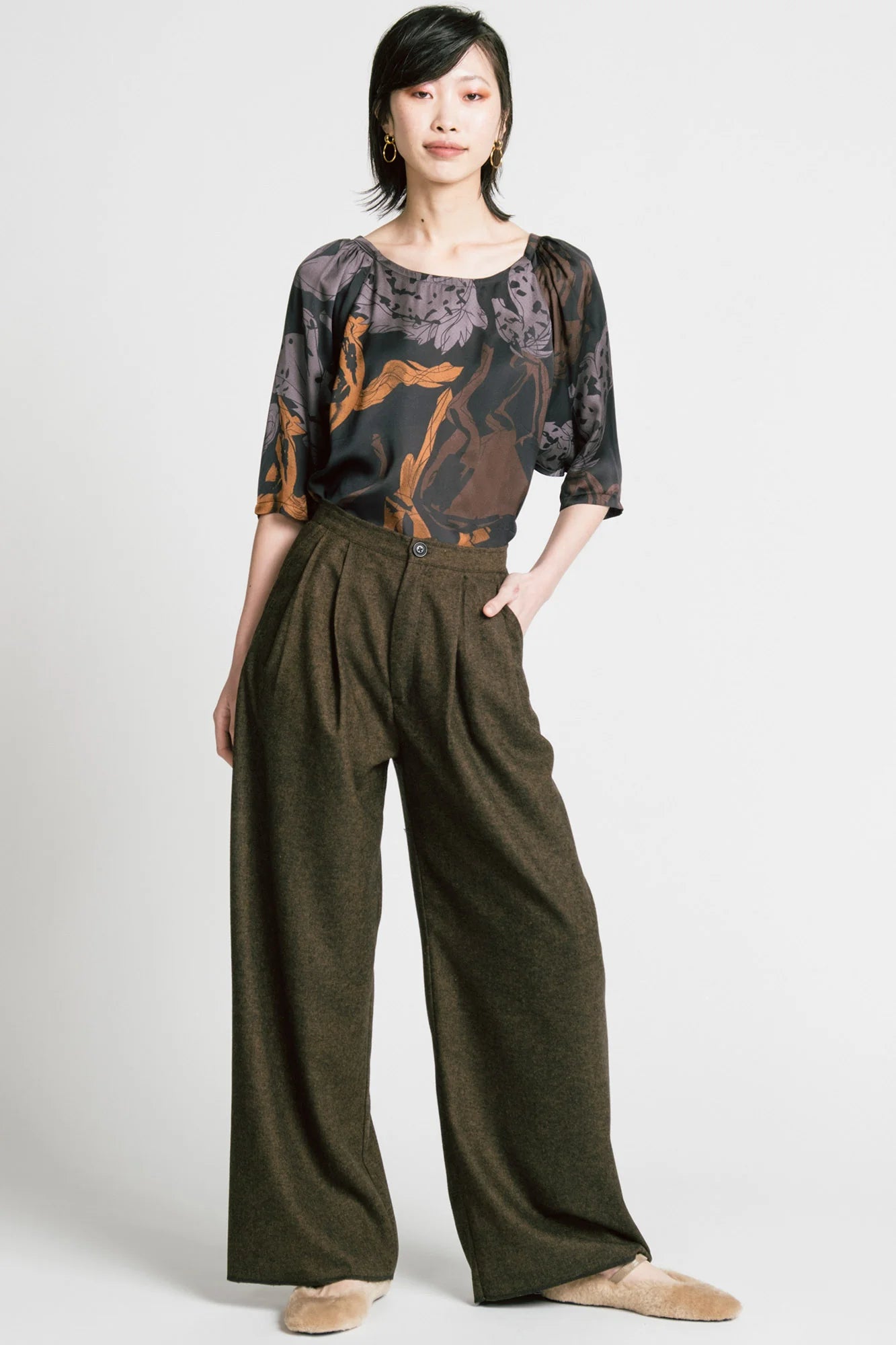A woman wearing the Memory Pant by Allison Wonderland in brown Herringbone print, wide-legged pants with front pleats, a zip and button closure, front pockets and back patch pockets. She is wearing them with the Flash Blouse in Horses print and standing in front of a white background. 