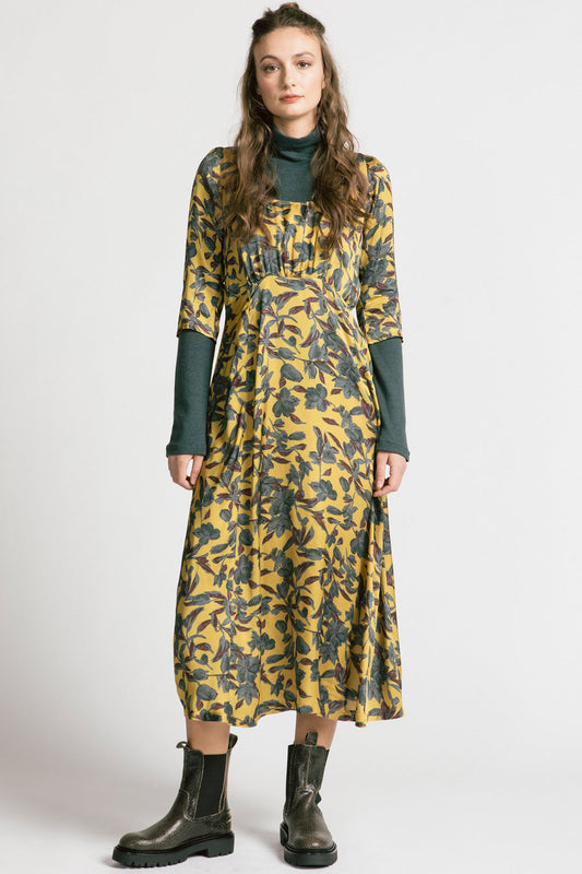 A woman wearing the Tempest Dress by Allison Wonderland in Gold Floral, with a  scoop neck, cinched empire waist, 3/4 sleeves, and long full skirt. She's wearing it over the Forever Turtleneck and is standing in front of a white background. 