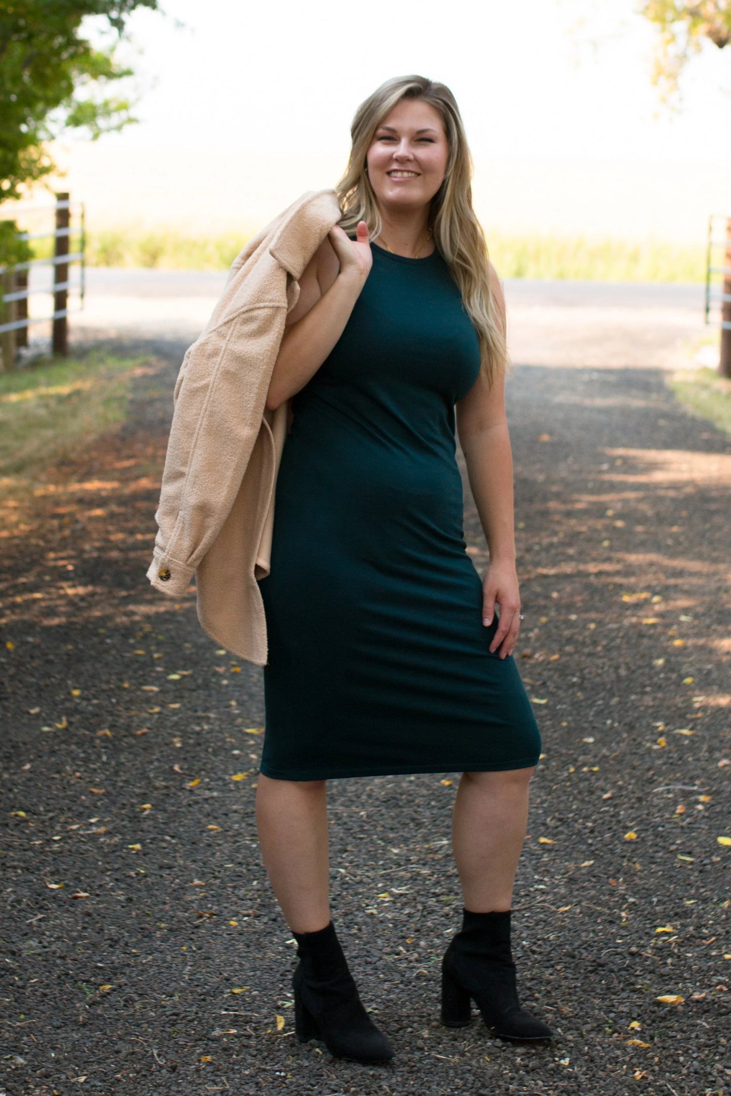 A woman wearing the Bev Tank Dress by Ang Hill in Juniper, a sleeveless, slim-fitting dress in cotton/modal with a high neck and a below the knee hem. She is wearing it with a beige jacket thrown over her shoulder and is standing outside on a gravel path. 
