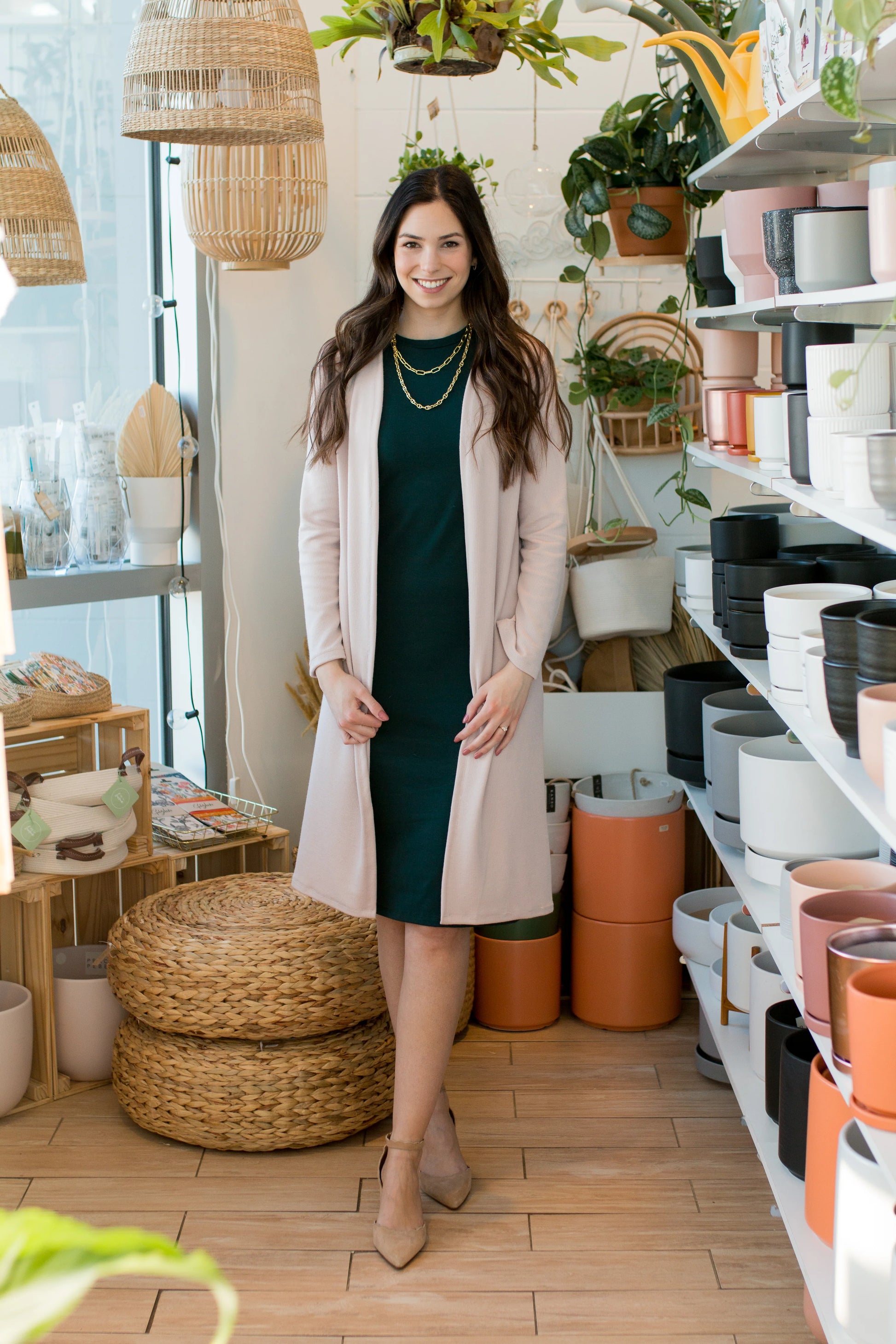 A woman wearing the Bev Tank Dress by Ang Hill in Juniper, a sleeveless, slim-fitting dress in cotton/modal with a high neck and a below the knee hem. She is wearing it with a beige sweater and standing in front of shelves of plants. 