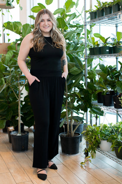 A woman wearing the Bree Wide Leg Pants by Ang Hill in Black, cropped wide-legged pants with a high elastic waist band and deep pockets. She is wearing them with a black tank top and standing in front of plants. 