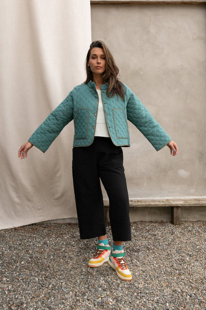 A woman wearing the Artemesia Jacket by Kazak in Mint, a quilted jacket with snaps up the front and large patch pockets. She is wearing it with a striped top and black pants and standing outside in front of a grey wall.
