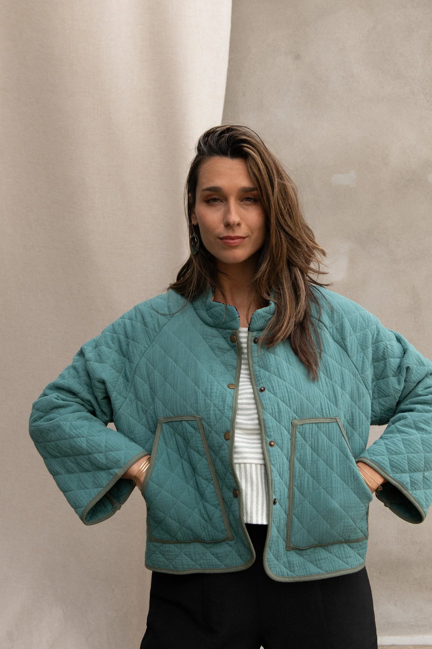 Waist up view of a woman wearing the Artemesia Jacket by Kazak in Mint, a quilted jacket with snaps up the front and large patch pockets. She is wearing it with a striped top and black pants and standing in front of a grey curtain. 